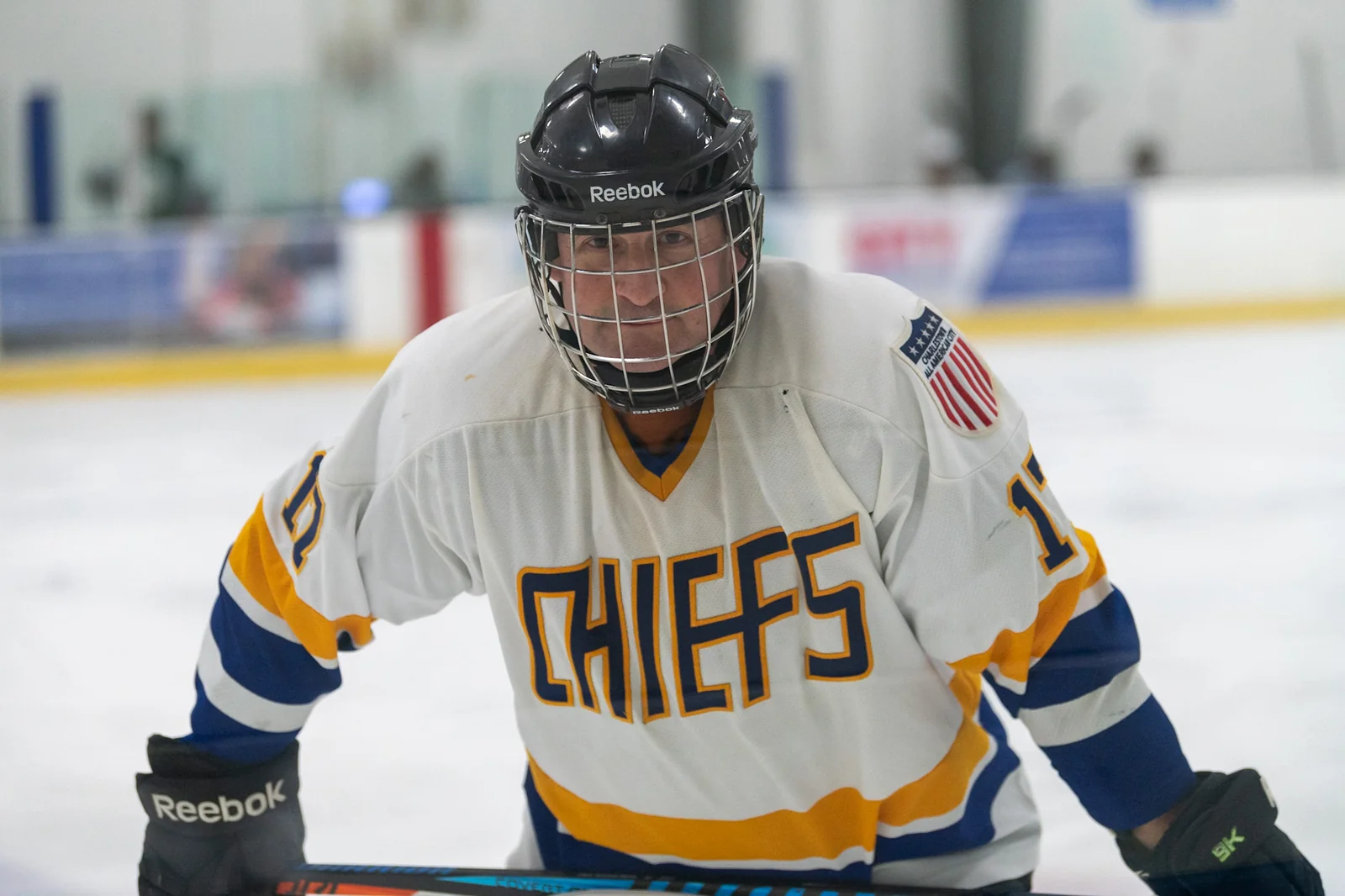 Scott Germaise on the ice at a rink, wearing a caged
                  hockey helmet and a white, blue, and gold jersey
                  lettered &lsquo;Chiefs&rsquo;, leaning toward the camera
                  at the boards.
