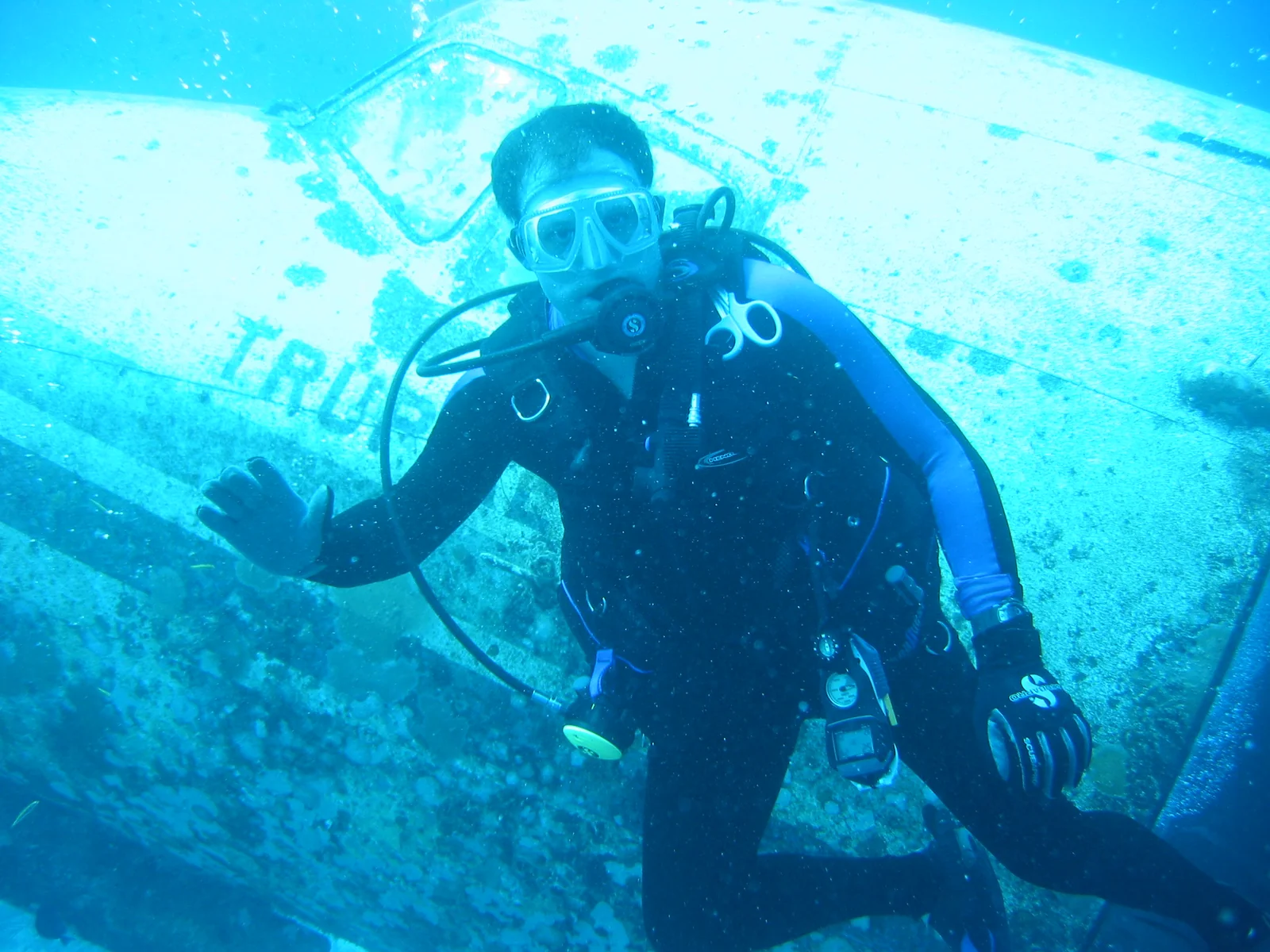Scott in scuba gear beside the barnacle-covered
                      fuselage of a sunken airplane, waving at the
                      camera.