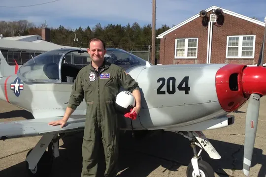 Scott in a flight suit beside a silver propeller
                      trainer aircraft marked 204.