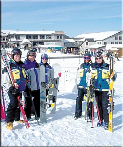 Five ski instructors in matching jackets posing with
                  skis and a snowboard at a base lodge on a sunny
                  day.