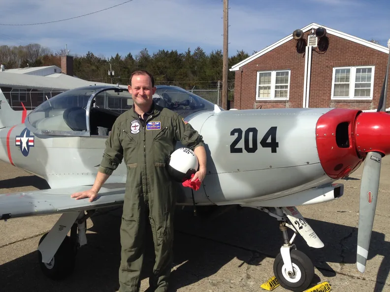 Scott in a flight suit beside a silver propeller
                      trainer aircraft marked 204.