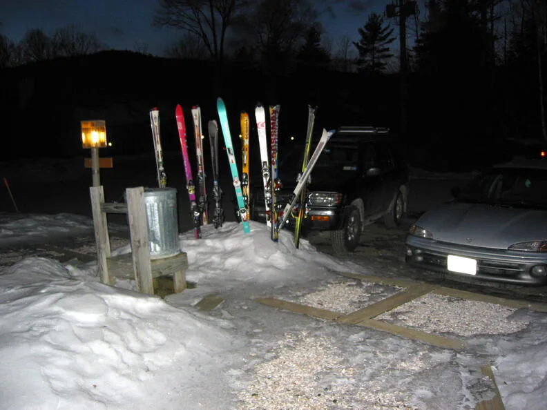 A row of skis standing upright in the snow in a
                      ski-area parking lot at dusk.
