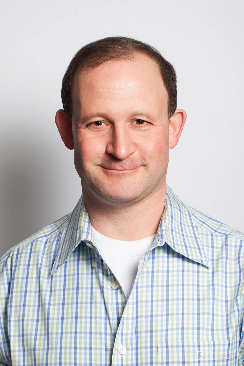 Scott Germaise, smiling, in a blue-and-green checked
                    shirt against a plain light studio background.